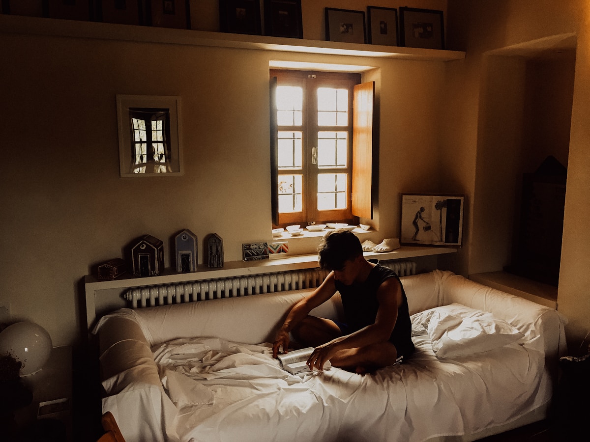 Man sitting on bed beside opened window in warm bedroom lighting